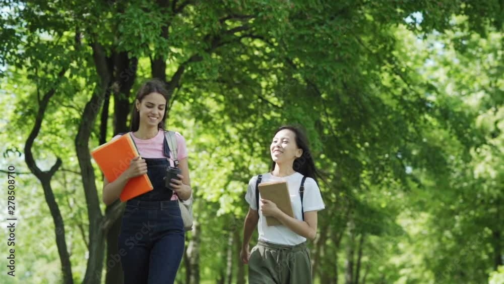 Panning slow motion shot of two happy female college friends with backpacks and textbooks walking through green park after classes and talking cheerfully on warm sunny day Panning slow motion shot of two happy female college friends with backpacks and textbooks walking through green park after classes and talking cheerfully on warm sunny day
