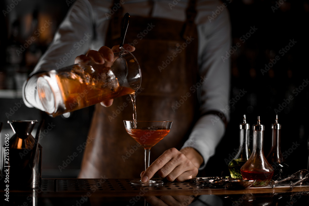 Professional male bartender pouring a brown alcoholic cocktail from the measuring cup to the glass in the dark