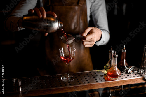 Bartender pouring a red alcoholic drink from the steel shaker through the sieve