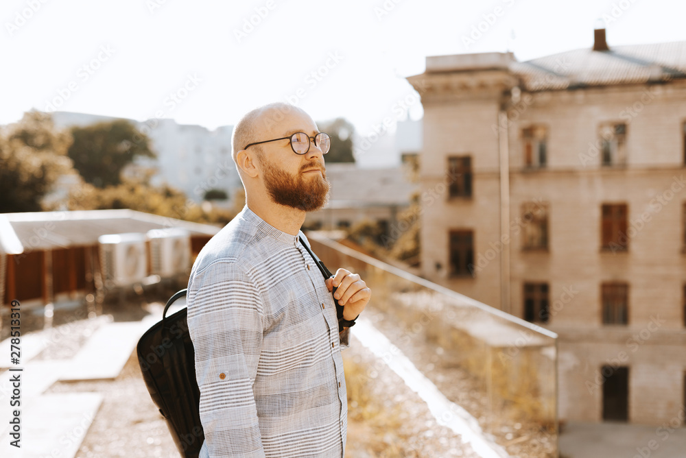 Photo of someone bearded guy,  standing on a terrace, wearing glasses, looking away and thinking