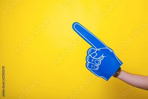 Fotografi Photo of a handsome arm,  wearing a big blue fan glove, over isolated yellow bac