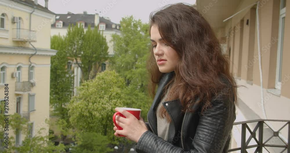 Young brunette girl in leather jacket standing on balcony enjoying the spectacular green city view and drinking hot tea. Young brunette girl in leather jacket standing on balcony enjoying the spectacular green city view and drinking hot tea.