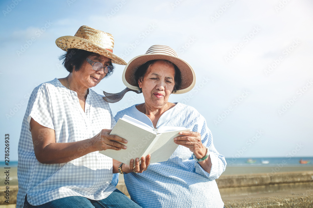 Two Asian elderly women sitting at the beach reading a book