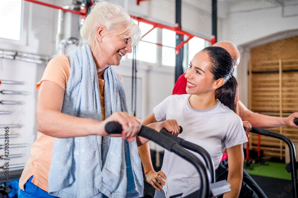 Obraz premium Senior woman biking at the gym with personal trainer