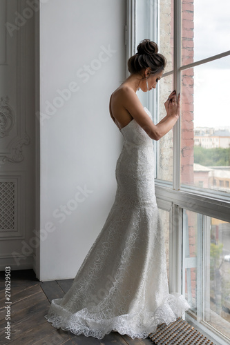 Beautiful wedding dress silhouette with a small train and embroidery. A young european bride stands alone near a panoramic window. Quarrel with the groom.