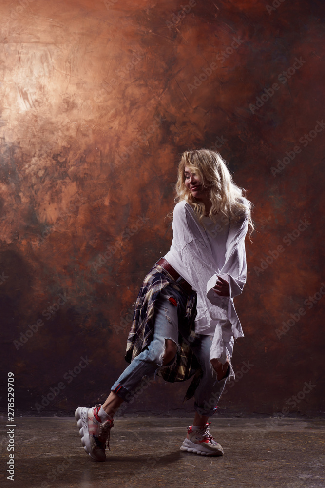 Photo of young dancing blonde girl in torn jeans on brown background