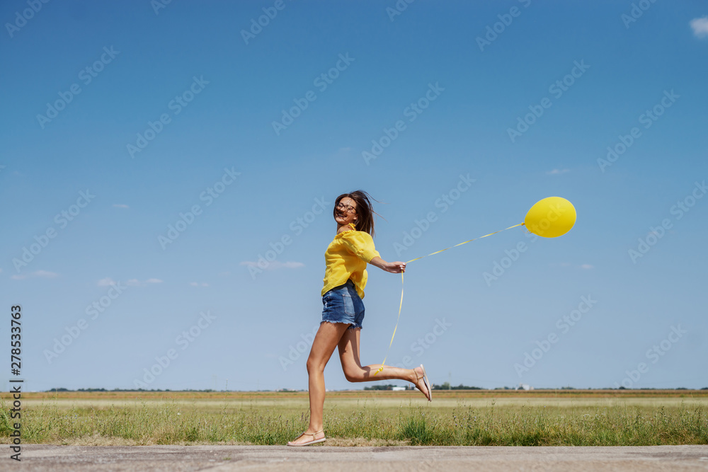 Gorgeous Caucasian teenage girl in yellow blouse, denim shorts and with eyeglasses running outdoor with yellow balloon in hand. Gorgeous Caucasian teenage girl in yellow blouse, denim shorts and with eyeglasses running outdoor with yellow balloon in hand.