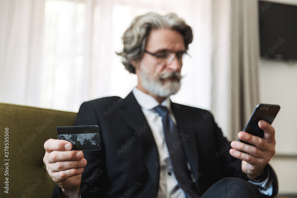 Handsome grey-haired senior businessman indoors at home dressed in formal clothes holding credit card using mobile phone. Handsome grey-haired senior businessman indoors at home dressed in formal clothes holding credit card using mobile phone.