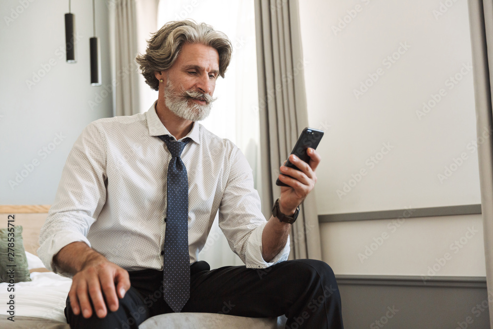 Pleased senior gray-haired business man sitting on bed indoors at home using mobile phone.