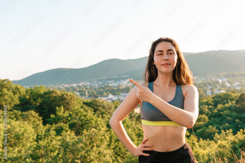 A young athletic woman points at something with her hand and looks at the camera. The concept of yoga, sports and meditation. In the background View of the village and the mountains. Copy space A young athletic woman points at something with her hand and looks at the camera. The concept of yoga, sports and meditation. In the background View of the village and the mountains. Copy space
