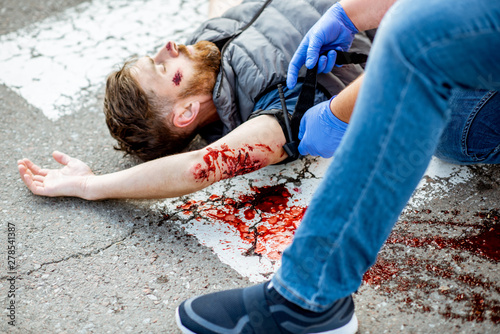 Applying first aid to the injured bleeding man, wearing tourniquet on the arm after the road accident on the pedestrian crossing photo