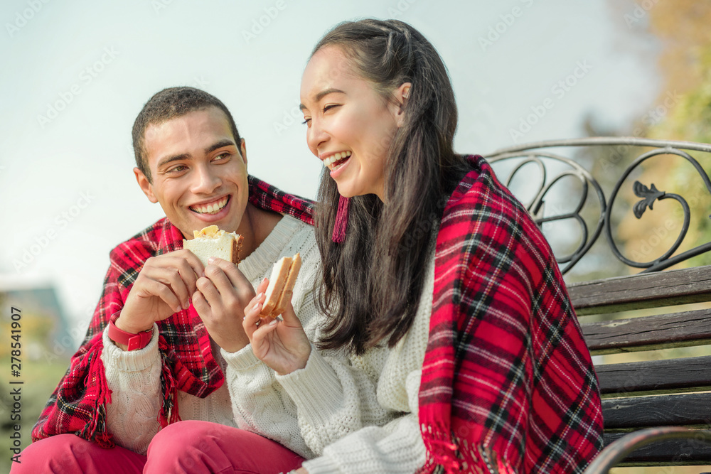 Joyful good-looking couple eating freshly-made sandwiches on a bench