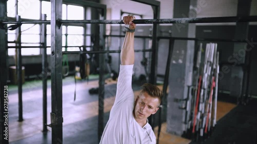 Man hangs on the bar in the gym on one hand. Young athlete in white T-shirt doing exercises on the bar, strengthening muscles and stretching. Lift and grip equipment for exercise at gym