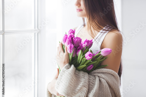 Attractive young woman with flowers indoors in the bedroom. Portrait of beautiful lady at home. Close up shot of female with tulips.