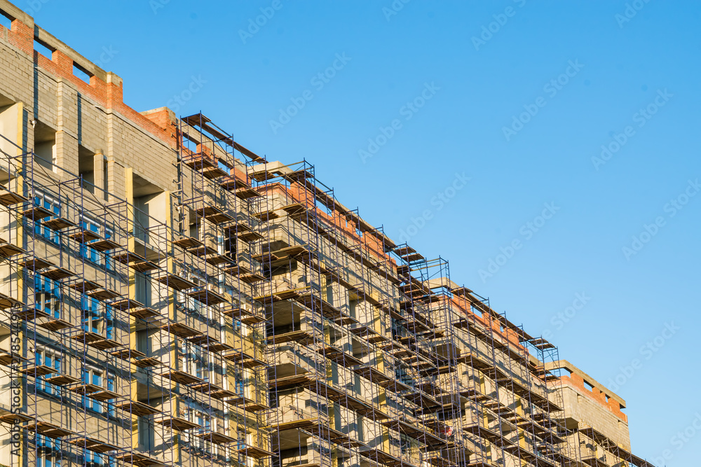Construction of a multi-storey residential building, a house in the scaffolding, illuminated by the evening sun against the blue sky Construction of a multi-storey residential building, a house in the scaffolding, illuminated by the evening sun against the blue sky
