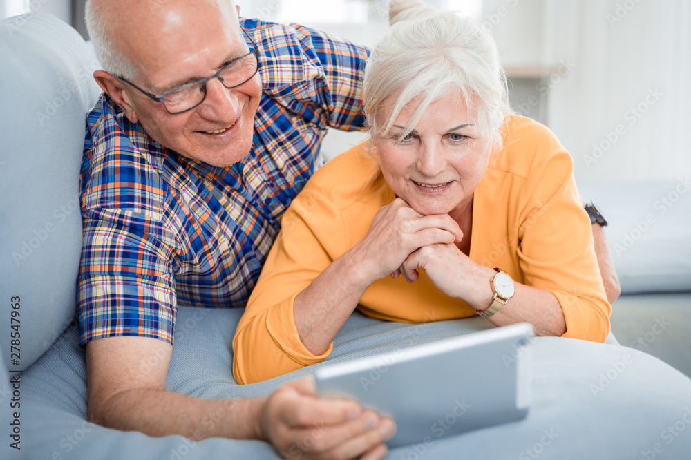 Happy senior couple looking at tablet lying on sofa at home