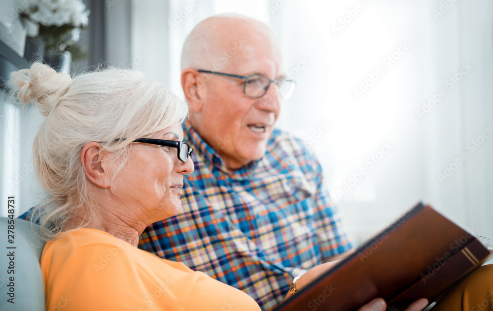 Cheerful senior couple holding family photo album sitting on sofa at home