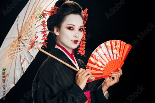 beautiful geisha in black kimono with red flowers in hair holding asian umbrella and hand fan isolated on black