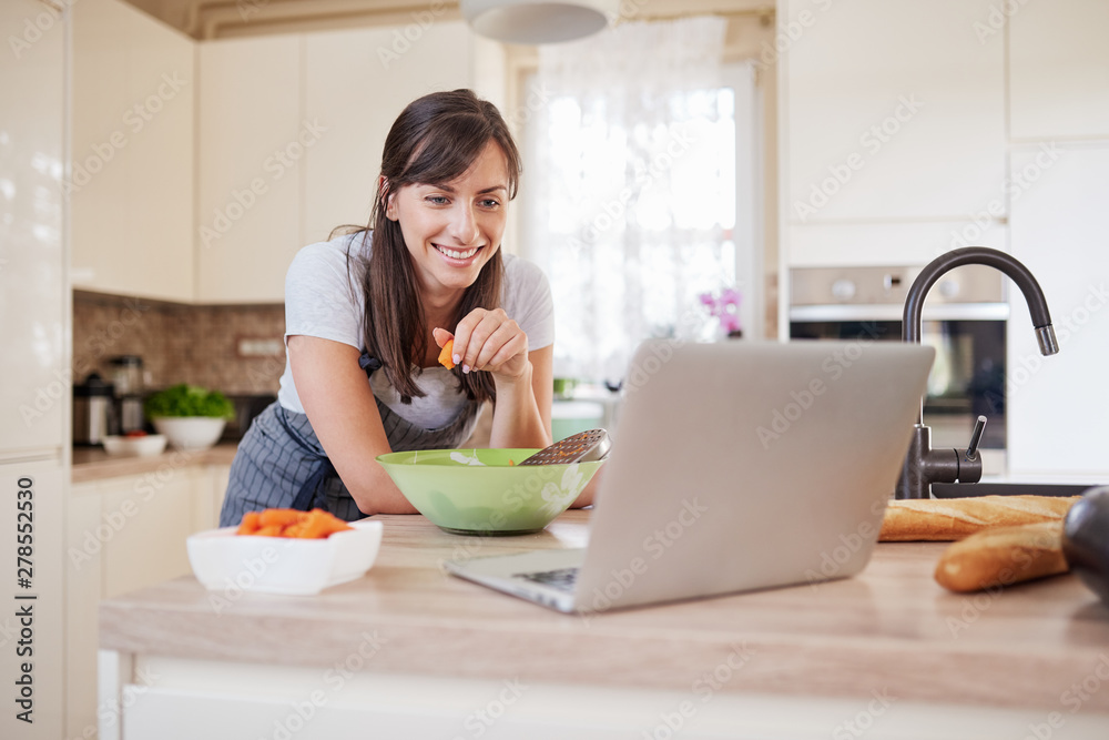 Pretty Caucasian brunette in apron leaning on kitchen counter and looking at laptop and following recipe for dinner. Woman holding carrot in hand.