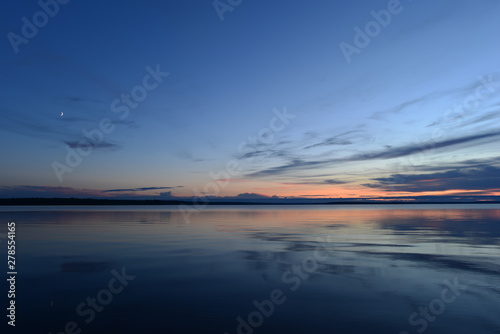 Moonrise in twilight light in the blue sky at sunset over the quiet mirror water of the lake