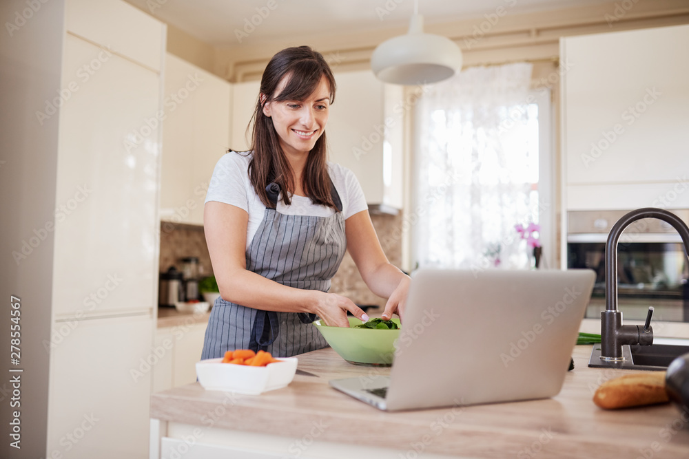 Beautiful Caucasian brunette in apron making vegetable salad for dinner or lunch and following recipe on laptop. Domestic kitchen interior. Beautiful Caucasian brunette in apron making vegetable salad for dinner or lunch and following recipe on laptop. Domestic kitchen interior.
