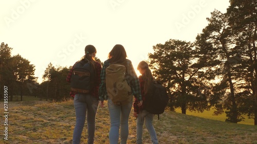 Hiker Girl. three girls travel  walk through woods to climb hill rejoice and raise their hands to top. girls travel with backpacks on country road. Happy family on vacation travels.