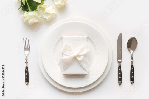 White table setting with bouquet rose, gift, dishware, silverware on white table. Romantic and wedding anniversary.