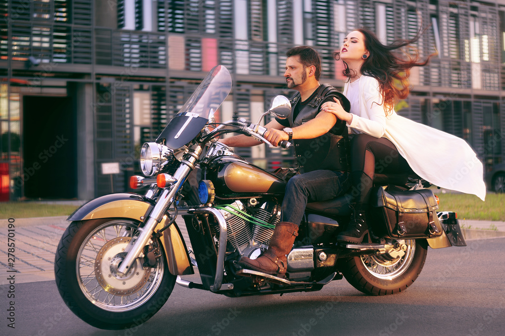 Couple on the background of the office on a motorcycle. The guy brought his girlfriend to work. Girl in a long white dress on a motorcycle. Couple against the backdrop of an industrial city.