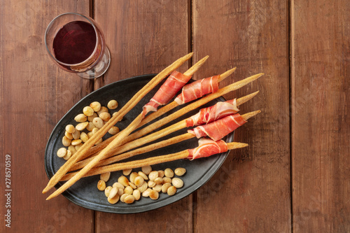 Prosciutto-wrapped Italian grissini with wine and roasted almonds, top view. Italian antipasti with parma ham, shot from the top on a dark rustic wooden background with a place for text