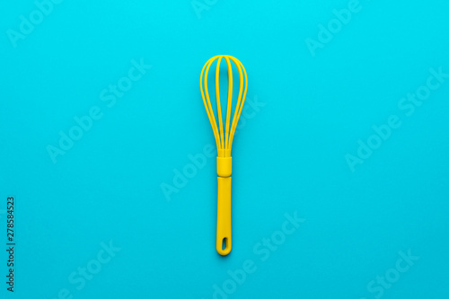Top view photo of vivid plastic kitchen utensil. Minimalist flat lay image of yellow whisk over turquoise blue background with copy space and central composition. Mixing kitchen tool on blue table.