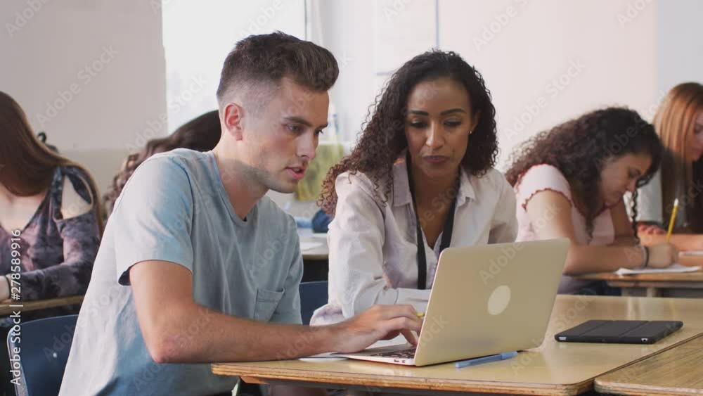 Female Tutor Giving One To One Support To Male Student Working At Desk On Laptop Female Tutor Giving One To One Support To Male Student Working At Desk On Laptop