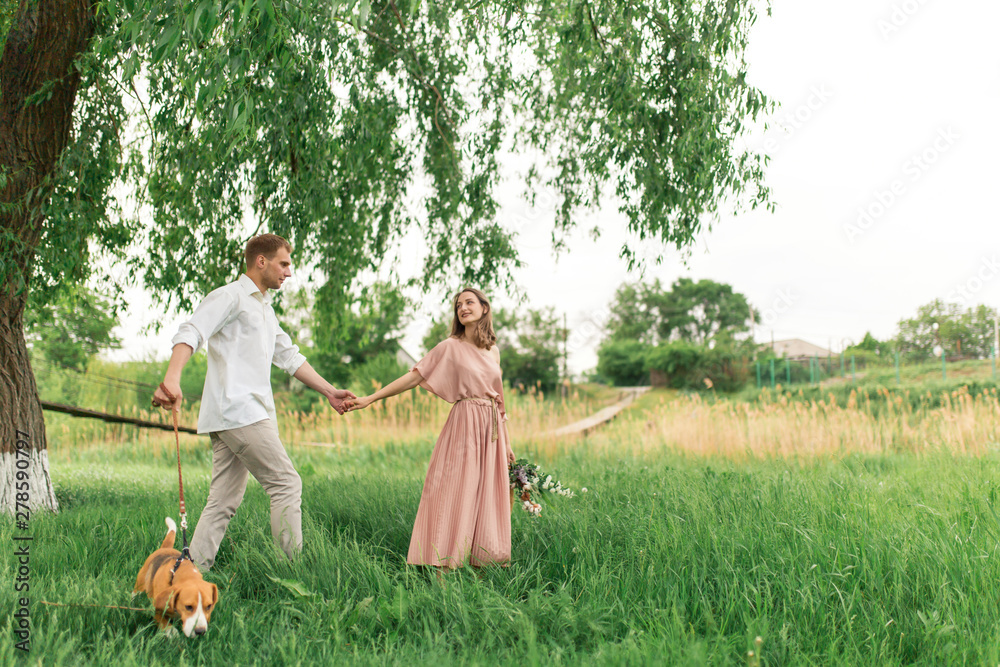 Obraz premium Young loving couple having fun and running on the green grass on the lawn with their beloved domestic dog breed Beagle and a bouquet of wildflowers