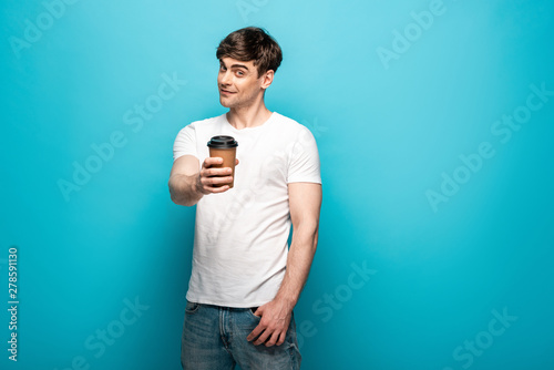 smiling young man holding paper cup in outstretched hand and looking at camera on blue background