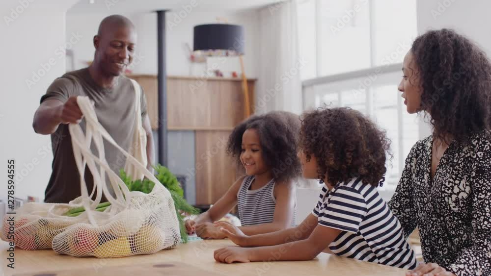 Father Returns Home From Shopping Trip As Mother Helps Children With Homework On Kitchen Table