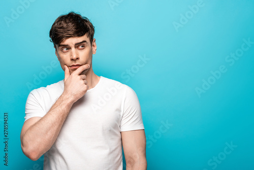 thoughtful young man holding hand near face while looking away on blue background