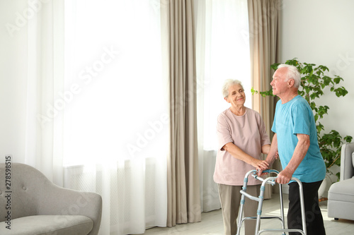 Elderly woman and her husband with walking frame indoors. Space for text