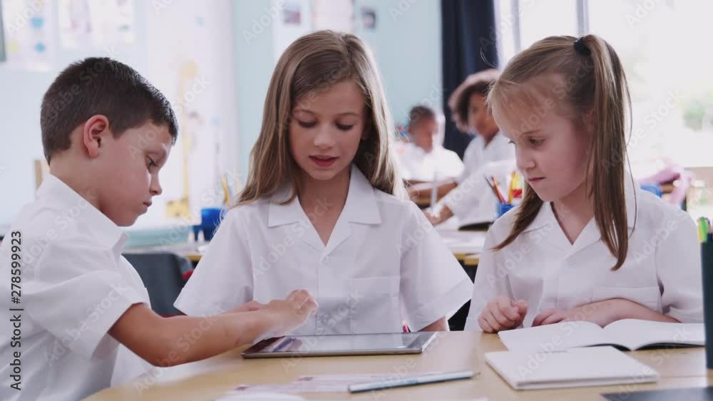 Three Elementary School Pupils Wearing Uniform Using Digital Tablet At Desk Three Elementary School Pupils Wearing Uniform Using Digital Tablet At Desk