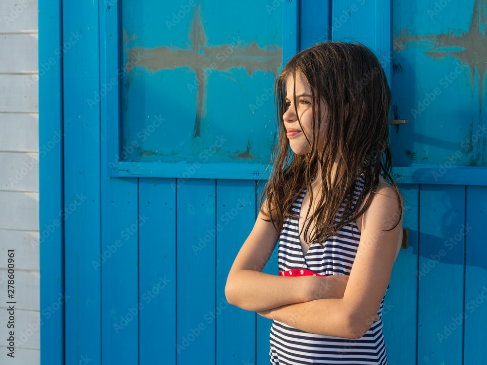 Fototapeta premium young beautiful girl with wet hair in a swimsuit posing on the beach against the background of a wooden blue house