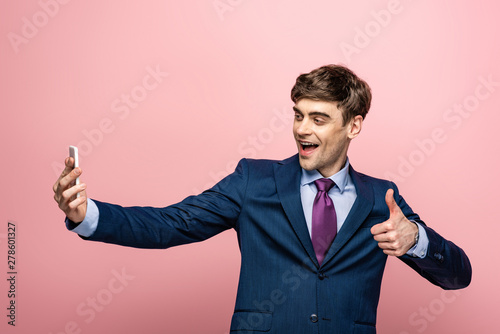 cheerful businessman showing thumb up while taking selfie with smartphone isolated on pink