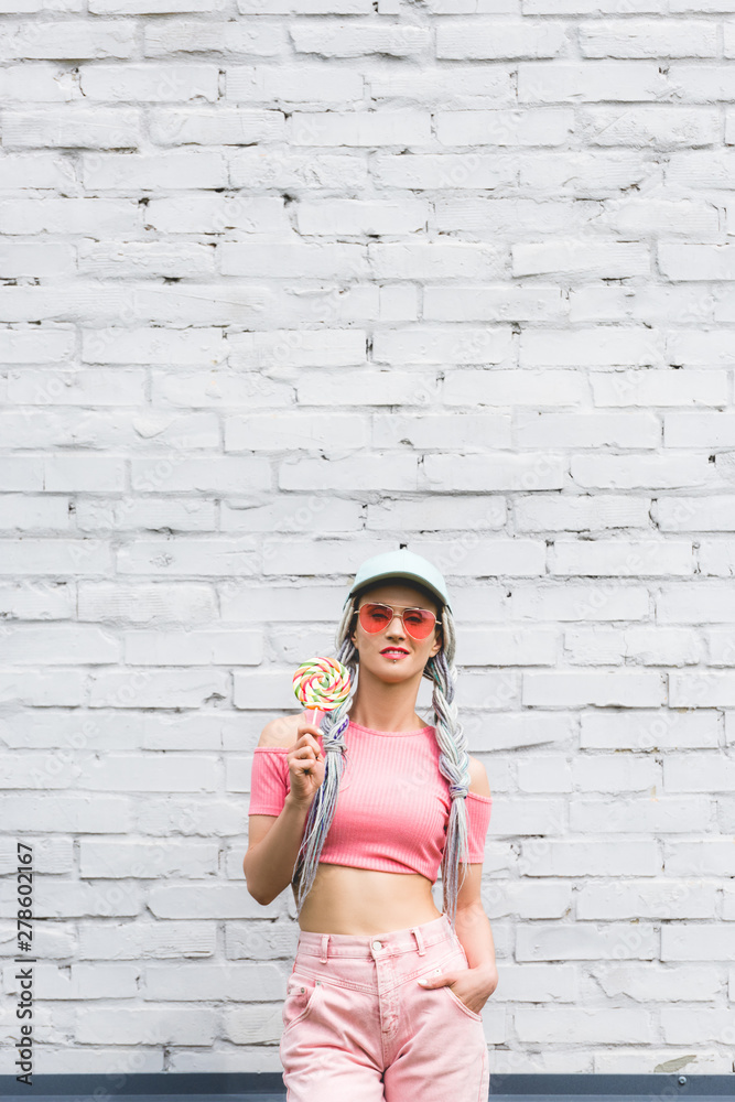 girl in hat and sunglasses holding lollipop near brick wall