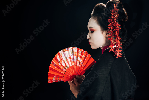 side view of beautiful geisha in black kimono with red flowers in hair holding traditional hand fan isolated on black photo