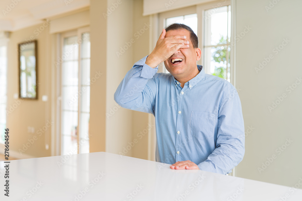 Middle age man sitting at home smiling and laughing with hand on face covering eyes for surprise. Blind concept.