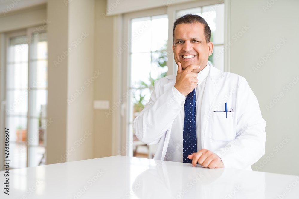 Middle age doctor man wearing medical coat at the clinic looking confident at the camera with smile with crossed arms and hand raised on chin. Thinking positive.