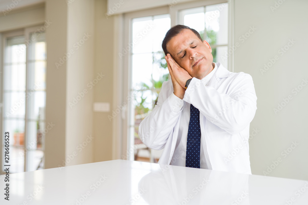 Middle age doctor man wearing medical coat at the clinic sleeping tired dreaming and posing with hands together while smiling with closed eyes.
