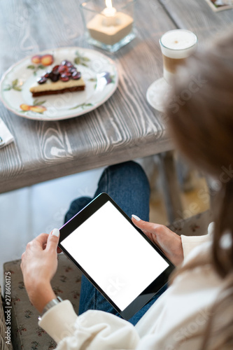 mock up tablet in hands of girl is her lap. Morning cafe, table with cake, coffee and lamp with burning candle. White screen. Copy space.