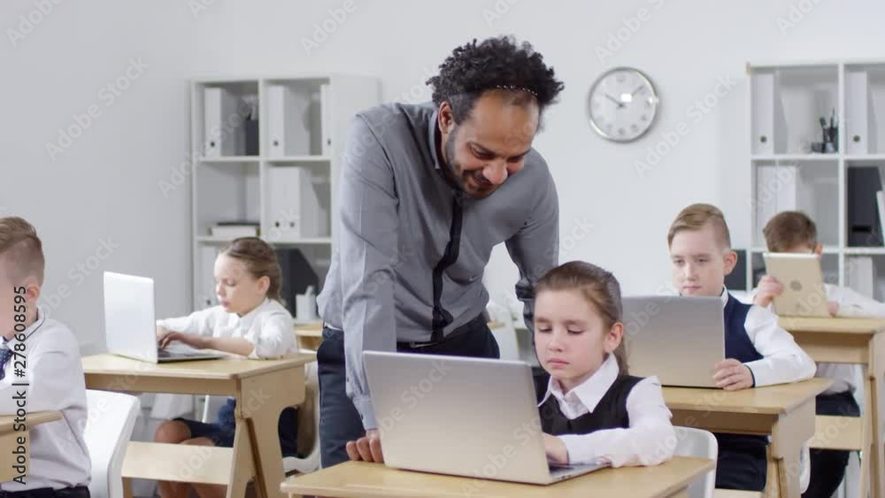Tracking shot of elementary school pupils sitting at desks with laptops during ICT class and working on assignment, and charismatic African American teacher standing next to schoolgirl and explaining Tracking shot of elementary school pupils sitting at desks with laptops during ICT class and working on assignment, and charismatic African American teacher standing next to schoolgirl and explaining