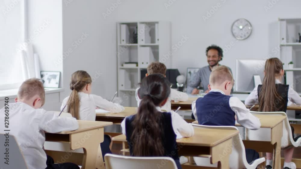 Full rear shot of group of pupils, wearing school uniform, sitting at desks in classroom and drawing on paper with graphite pencils, and male African American teacher at table supervising themFull rea