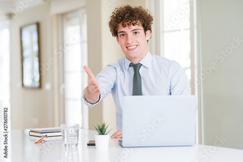 Young business man working with computer laptop at the office smiling friendly offering handshake as greeting and welcoming. Successful business.