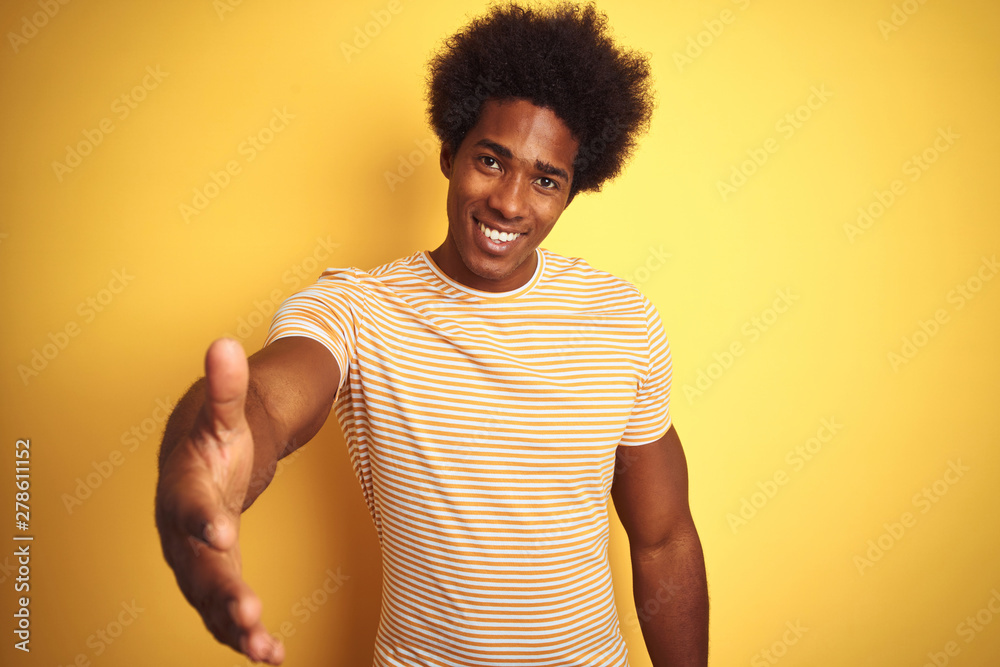 American man with afro hair wearing striped t-shirt standing over isolated yellow background smiling friendly offering handshake as greeting and welcoming. Successful business. American man with afro hair wearing striped t-shirt standing over isolated yellow background smiling friendly offering handshake as greeting and welcoming. Successful business.