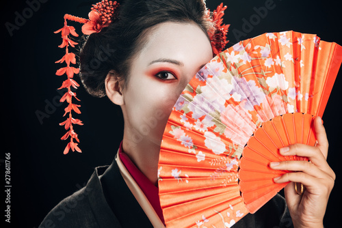 beautiful geisha with red flowers in hair holding traditional hand fan isolated on black